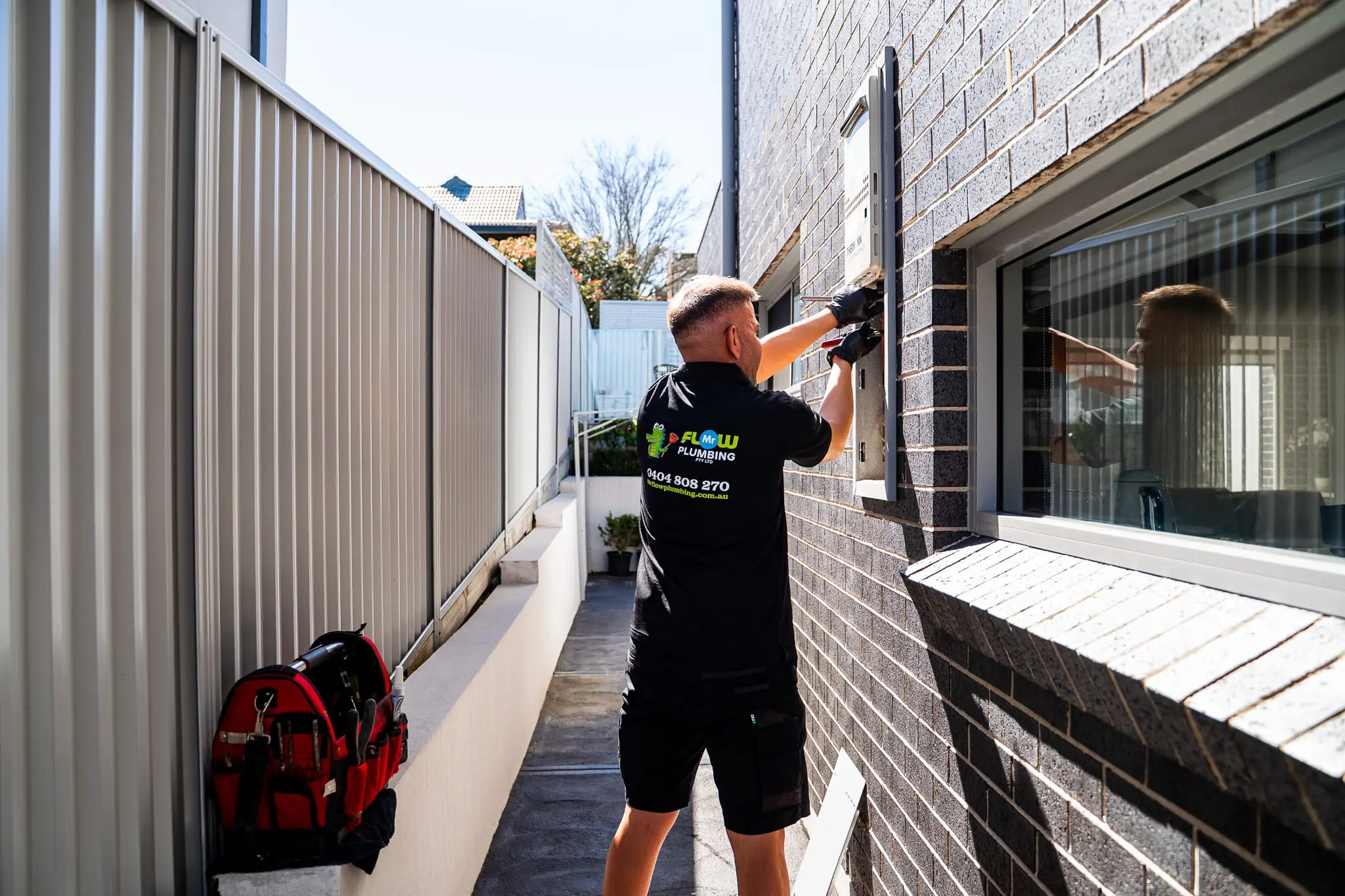 Mr Flow plumber at a continuous-flow hot water unit in a side passage